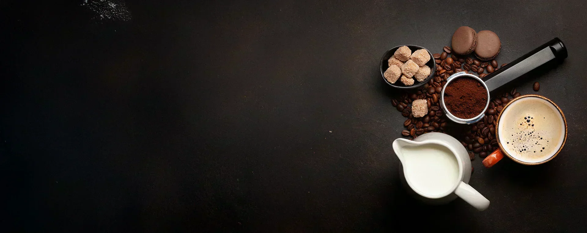 A coffee setup with ground coffee, beans, cream, and sweets on a dark background, showcasing coffee brewing essentials.
